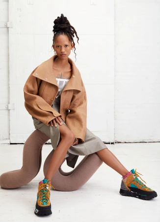 A woman with a high bun and statement earrings wears a short brown jacket, skirt, and high-top hiking boots while seated indoors.