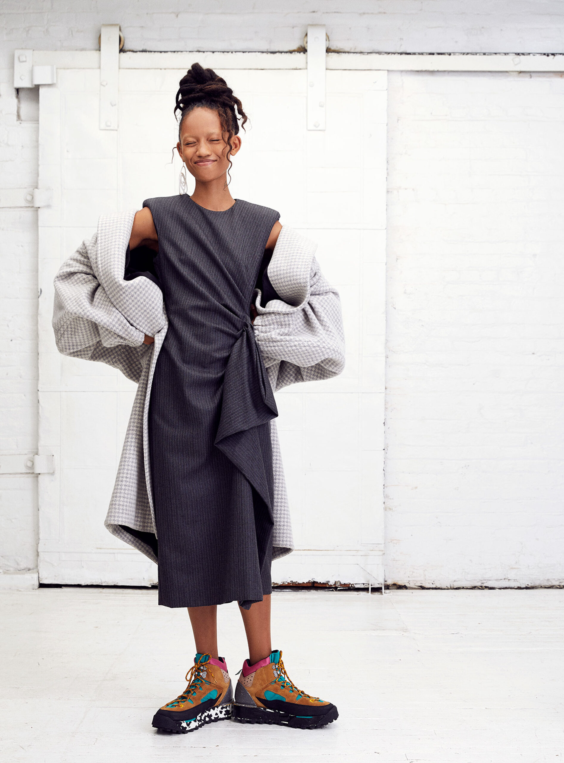 A slender woman with dark hair in an updo poses in a gray dress and textured coat in front of a white, sliding barn door.