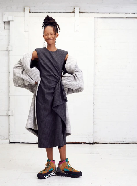 A slender woman with dark hair in an updo poses in a gray dress and textured coat in front of a white, sliding barn door.