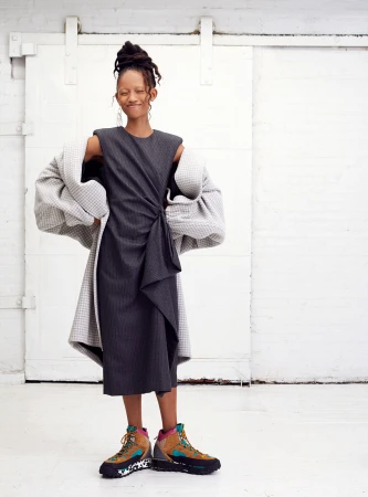 A slender woman with dark hair in an updo poses in a gray dress and textured coat in front of a white, sliding barn door.