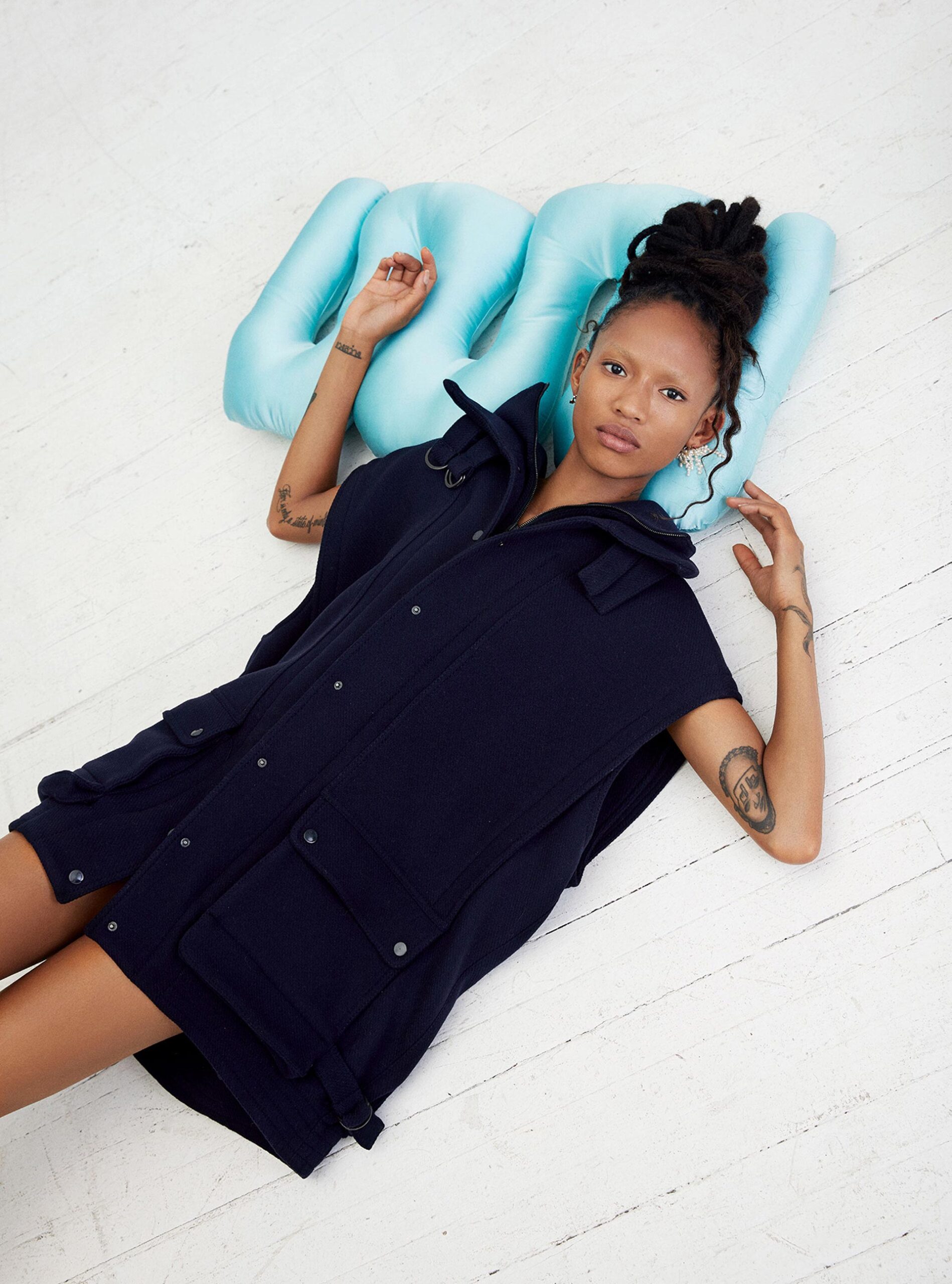 A young adult woman poses in a black vest and sits on a light blue pillow on a distressed white wood floor in a studio setting.