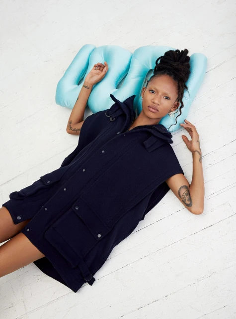 A young adult woman poses in a black vest and sits on a light blue pillow on a distressed white wood floor in a studio setting.