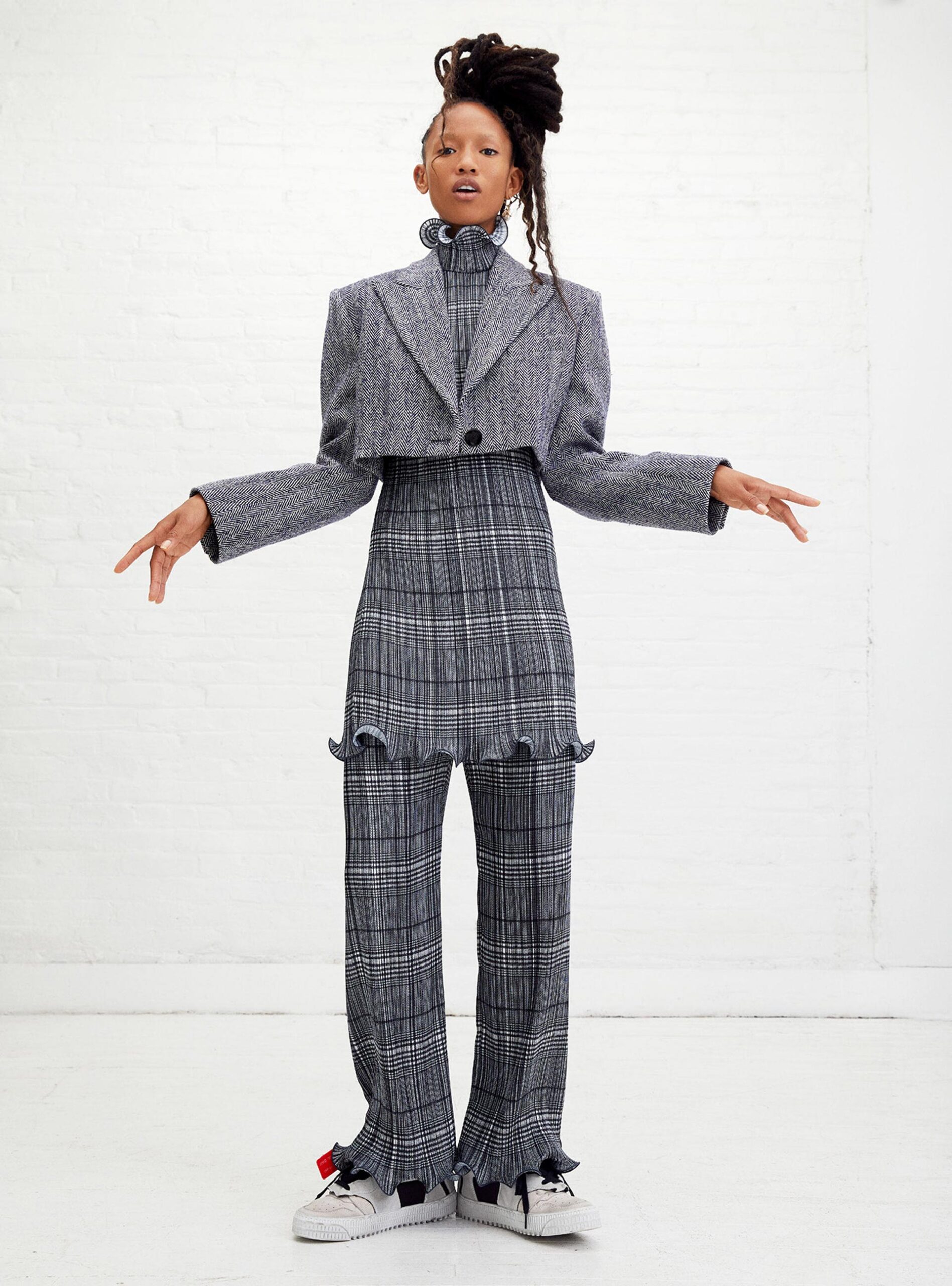 A woman poses in grey patterned separates consisting of a cropped blazer and pants with frills. She has long dreadlocks.