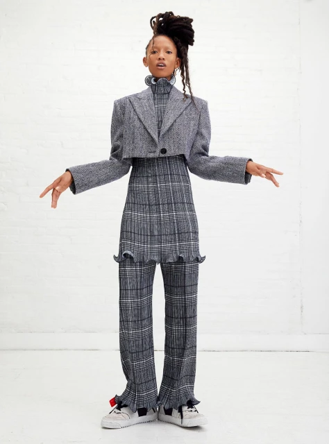 A woman poses in grey patterned separates consisting of a cropped blazer and pants with frills. She has long dreadlocks.