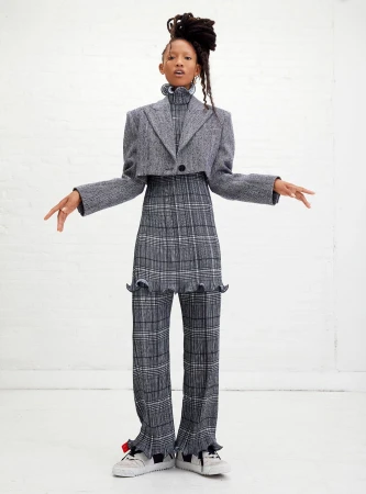 A woman poses in grey patterned separates consisting of a cropped blazer and pants with frills. She has long dreadlocks.