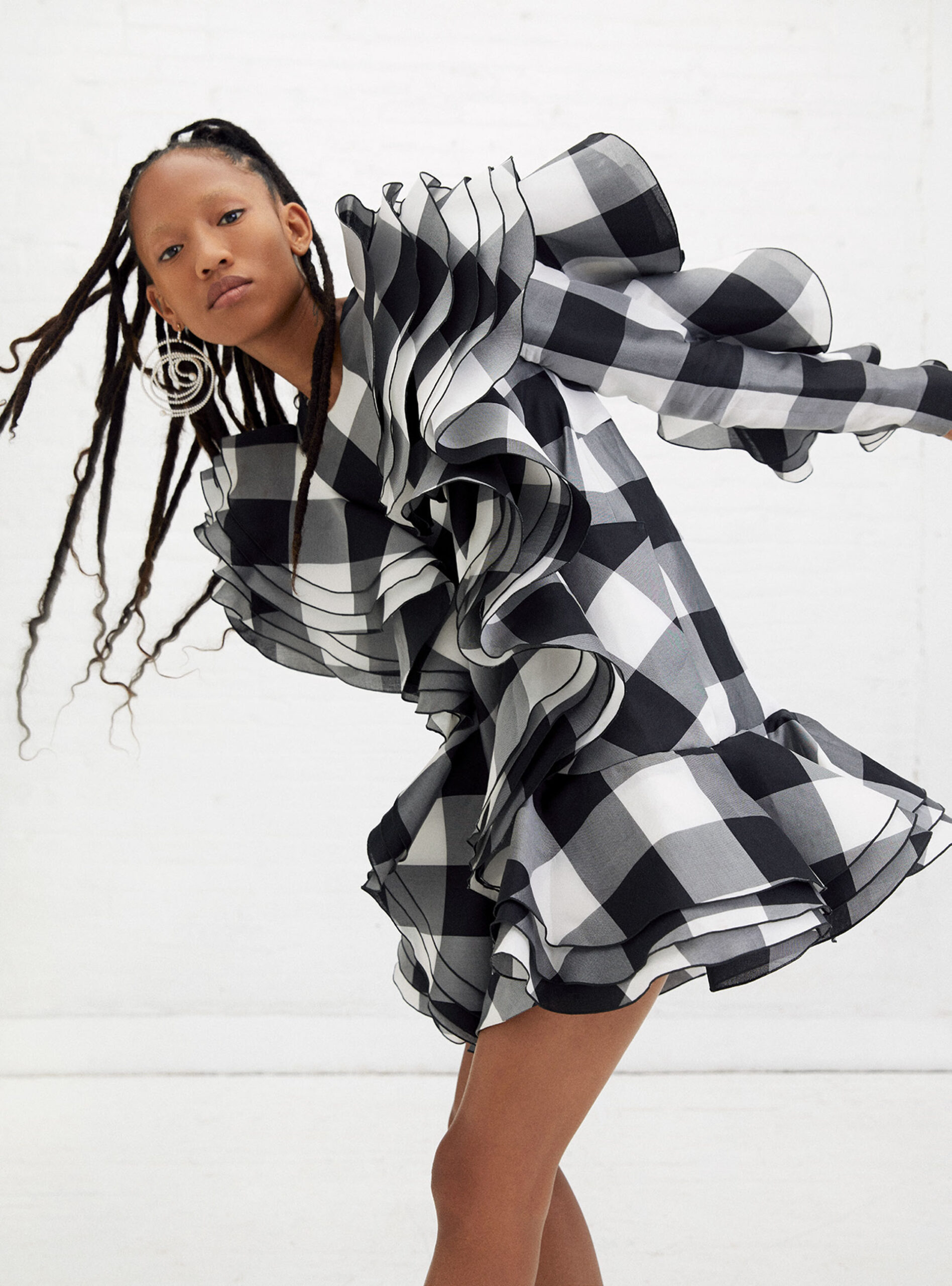 Fashion editorial shot of a woman in a short black and white gingham dress with oversized ruffles, set against a bright white backdrop.
