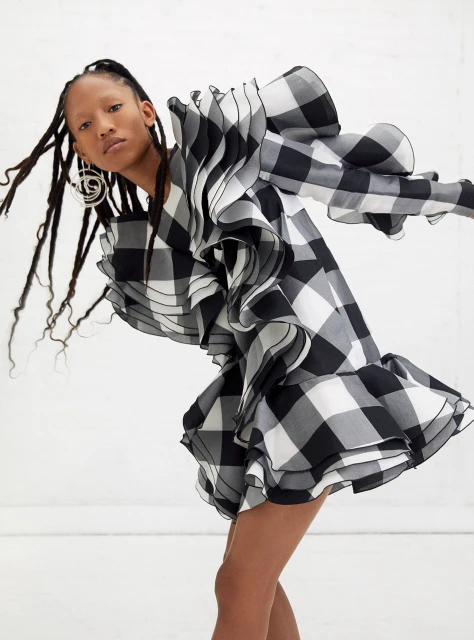Fashion editorial shot of a woman in a short black and white gingham dress with oversized ruffles, set against a bright white backdrop.