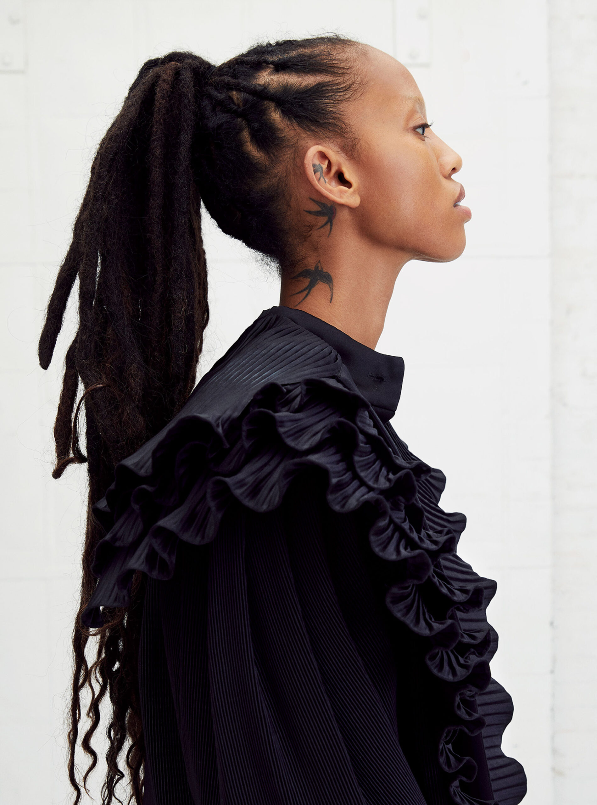 Side profile portrait of a slender Black woman with dreadlocks pulled into a high ponytail, wearing a ruffled black top and a bird tattoo.