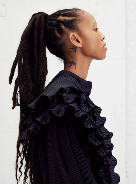 Side profile portrait of a slender Black woman with dreadlocks pulled into a high ponytail, wearing a ruffled black top and a bird tattoo.