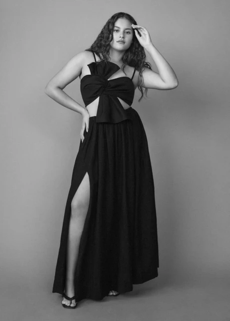 Full-length portrait in black and white of a woman in a black two-piece outfit with a high slit, standing in front of a grey backdrop.