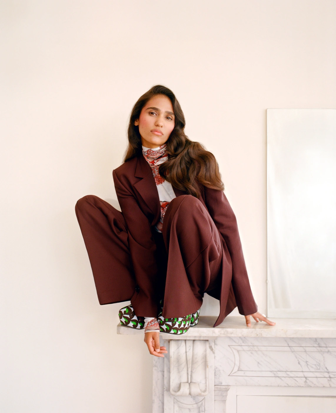 Woman wearing a brown suit and patterned top crouching on a marble mantelpiece in a room