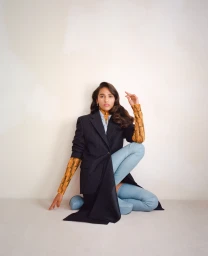 Woman in dark pinstripe blazer and light blue leather trousers kneeling against a seamless neutral studio backdrop.
