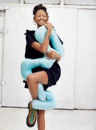 Smiling woman in dark clothes hugging a large light blue curved cushion against a white paneled wall