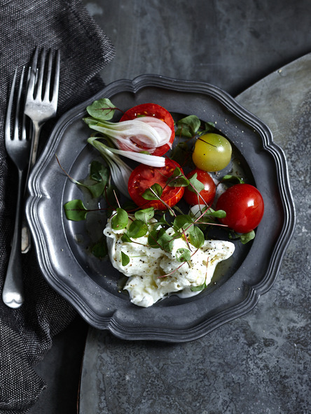 Overhead shot of a pewter plate with halved red tomatoes, burrata cheese, and microgreens next to two forks, on a dark background.
