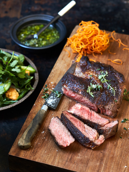 Close-up shot of a juicy steak, artfully sliced and arranged on a rustic wooden board, accompanied by a vibrant green salad and sauce.