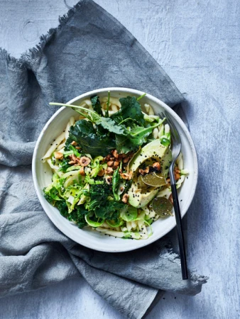 Overhead shot of a bowl of udon noodles with avocado, kale, sprouts, and nuts, sitting on a gray textured cloth.