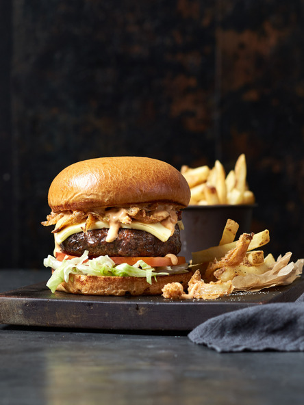 Burger with cheese, lettuce, tomato, and fries, served on a wooden board against a dark background in a food photography style.