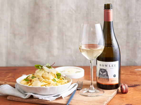 Overhead shot of white wine in a glass with pasta topped with shaved parmesan, a wine bottle, on a wooden board.