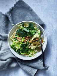 Bowl of pasta salad topped with avocado slices, greens, and chopped nuts next to a dark fork on a grey textile
