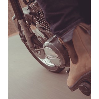 Close-up of a motorcyclist's foot on a vintage motorcycle, showcasing the engine and wheel. The focus is on the motorcycle's details.