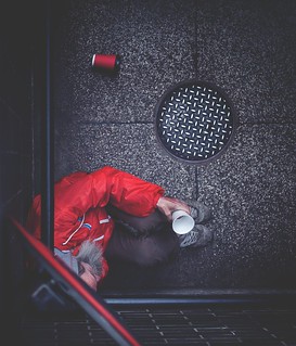 Adult sits on a sidewalk with a red cup and a white coffee cup, next to a sewer grate and with a discarded can in the background.