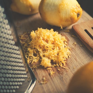 Overhead shot of a cutting board with lemon zest, whole lemons, grater, and wooden utensil featuring bright yellow tones and a rustic, culinary theme.