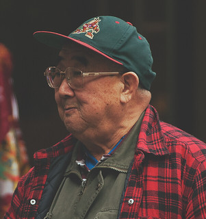 Elderly man in glasses and a cap smiles while wearing a red-and-black flannel shirt, photographed outdoors in natural light.