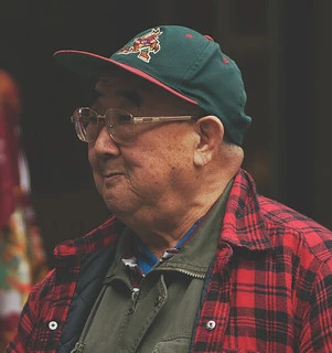 Elderly man in glasses and a cap smiles while wearing a red-and-black flannel shirt, photographed outdoors in natural light.