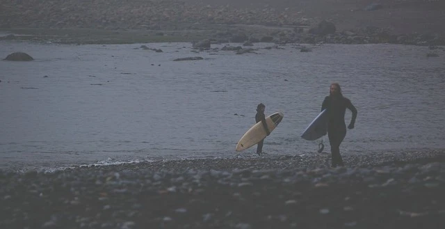 Two surfers in wetsuits walk on a rocky beach carrying surfboards near calm, gray water and an overcast sky at dusk or dawn.