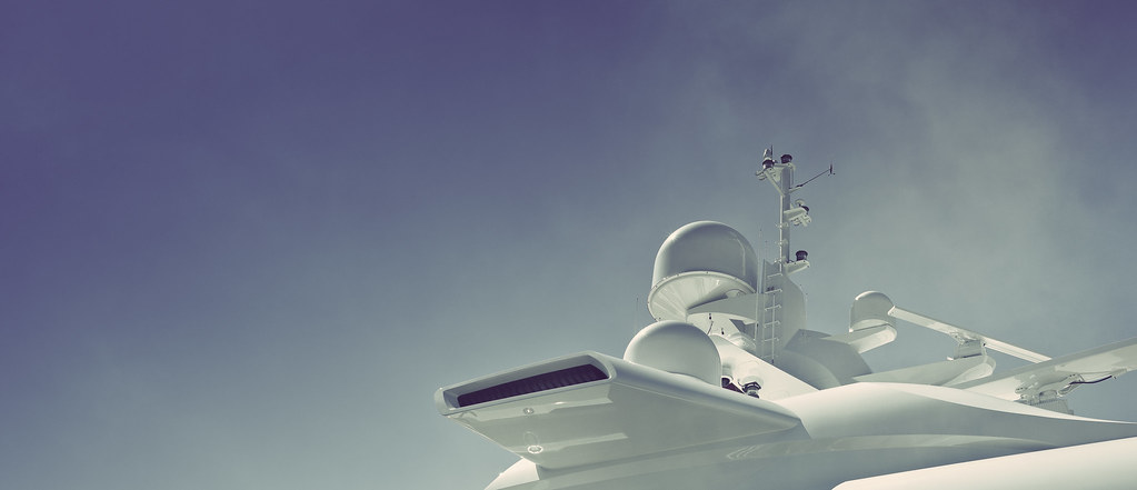 Low-angle shot of a modern, sleek white yacht featuring prominent antennas and radar dome against a soft, gradient blue sky backdrop.