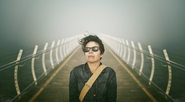 Woman in sunglasses stands on a foggy bridge. She wears a denim jacket and a crossbody bag with her hair blowing in the wind.