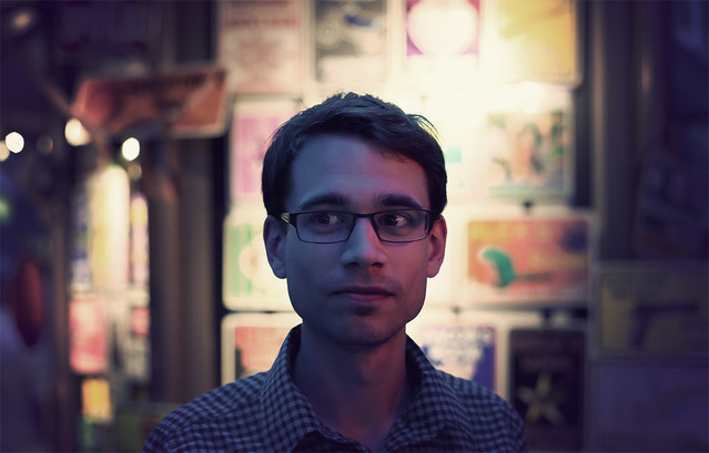 Adult male with glasses and plaid shirt looks to the side against a backdrop of colorful posters in an indoor setting with purple light.