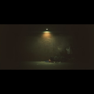 Wide shot of an adult male laying on the ground in an indoor setting with a concrete wall and dim lighting overhead.