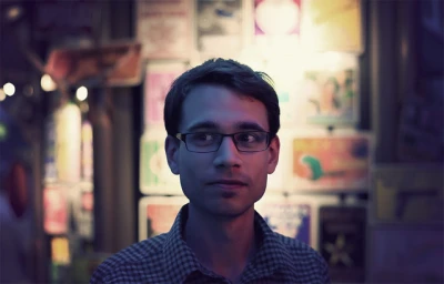 Portrait of a man wearing glasses and a plaid shirt against a softly lit background with posters