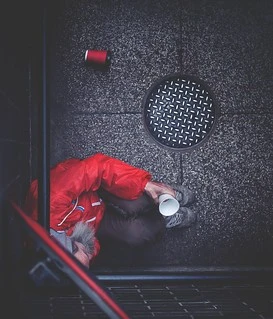 High-angle shot of a person in a red jacket seated on speckled grey pavement next to a round metal manhole cover.