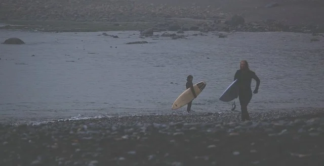 Two surfers in black wetsuits carrying surfboards along a rocky beach under an overcast sky.