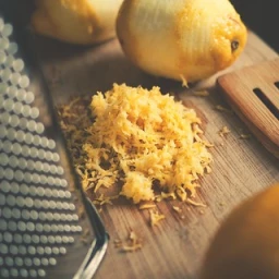 Zested lemon peel and a whole lemon on a wooden cutting board next to a metal grater