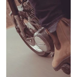 Close-up shot of a motorcycle engine and front wheel with a rider wearing brown boots and dark jeans.
