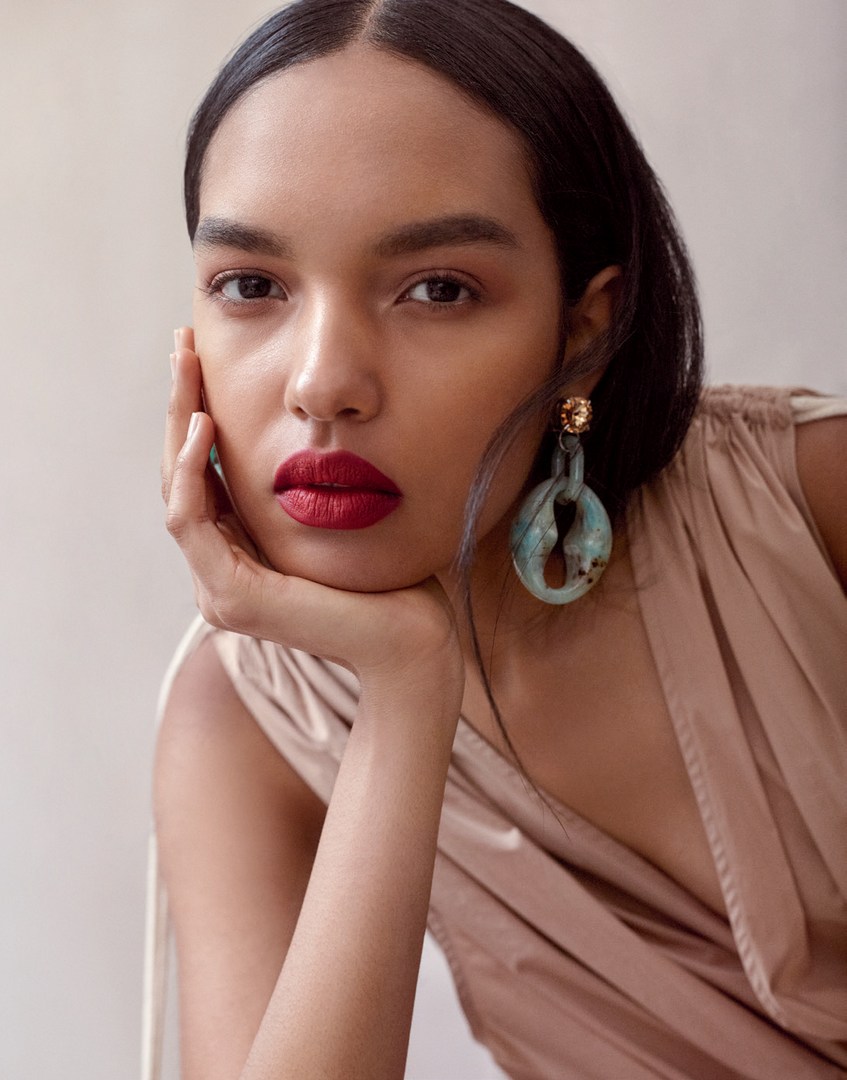 Close-up portrait of a woman with bold red lipstick and chunky blue resin earrings wearing a beige top.