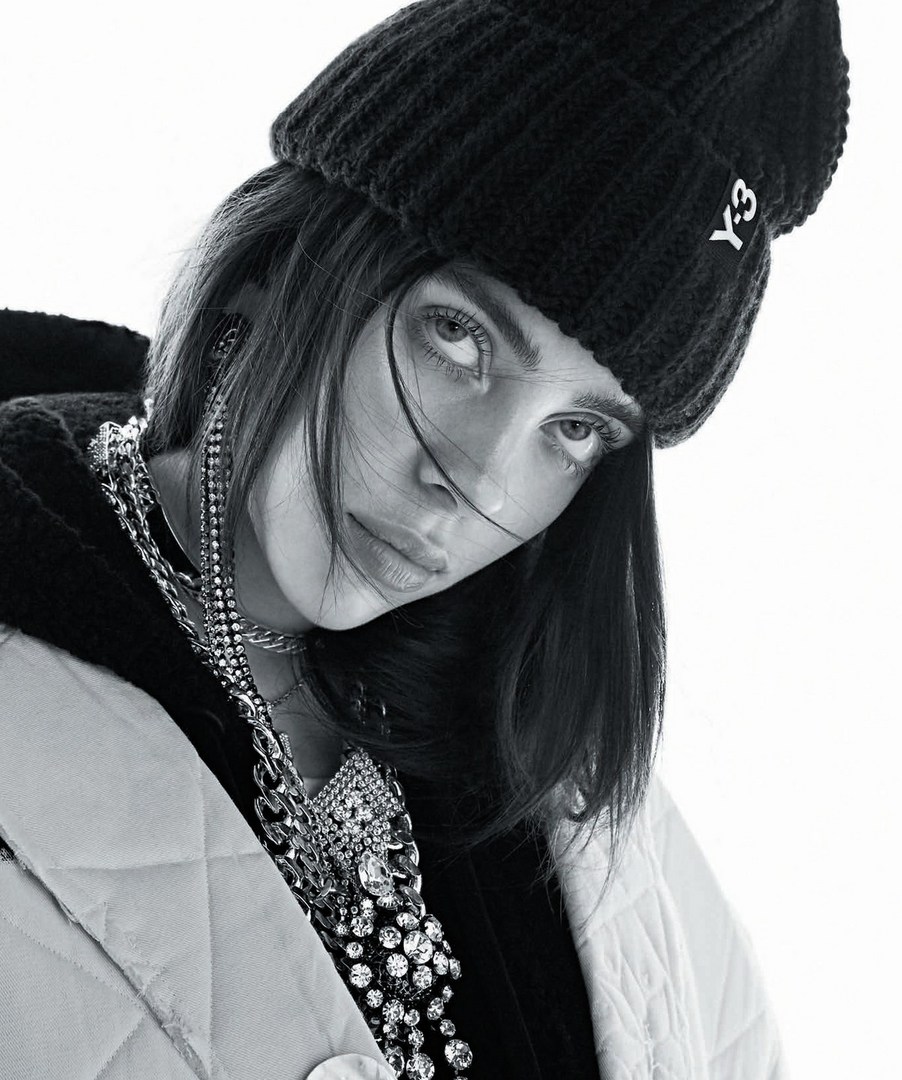 Black and white portrait of a woman wearing a black beanie, quilted jacket, and large chain necklace looking at the camera indoors.