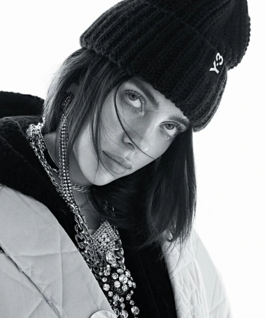 Black and white portrait of a woman wearing a black beanie, quilted jacket, and large chain necklace looking at the camera indoors.