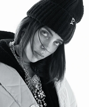 Black and white portrait of a woman wearing a black beanie, quilted jacket, and large chain necklace looking at the camera indoors.