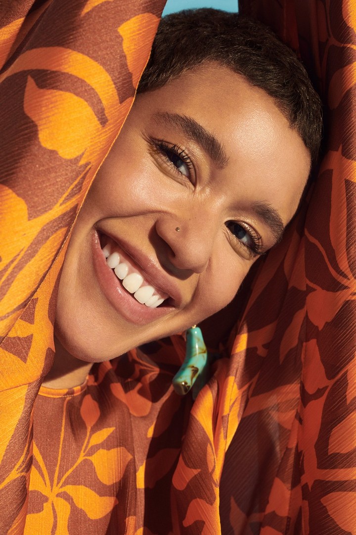 Close-up of a smiling young woman with short hair wearing patterned top and earrings, set against a bright blue sky background.