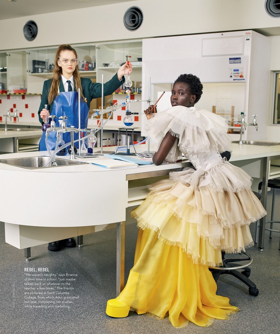 Editorial fashion shot in a science lab featuring two women: one in a lab coat working with beakers, the other in a formal yellow gown.