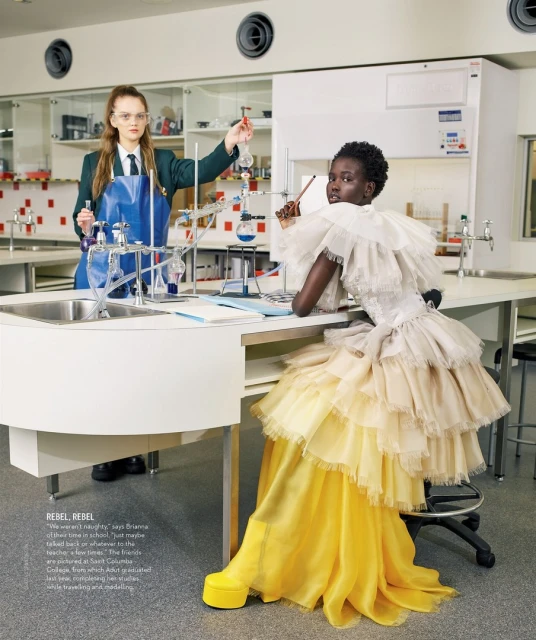 Editorial fashion shot in a science lab featuring two women: one in a lab coat working with beakers, the other in a formal yellow gown.