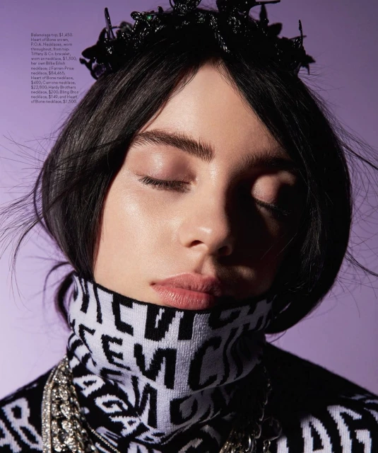 Close-up of a woman with her eyes closed, wearing a black and white turtleneck sweater and multiple silver necklaces, against purple backdrop.