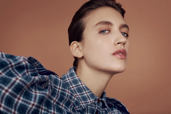 Close-up portrait of a woman with slicked-back brown hair wearing a blue plaid shirt against a warm brown background.