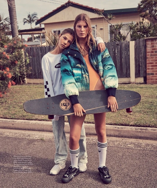 Two fashion models pose outdoors in a suburban street, one holding a skateboard and wearing a puffer jacket, the other in a long-sleeve shirt.