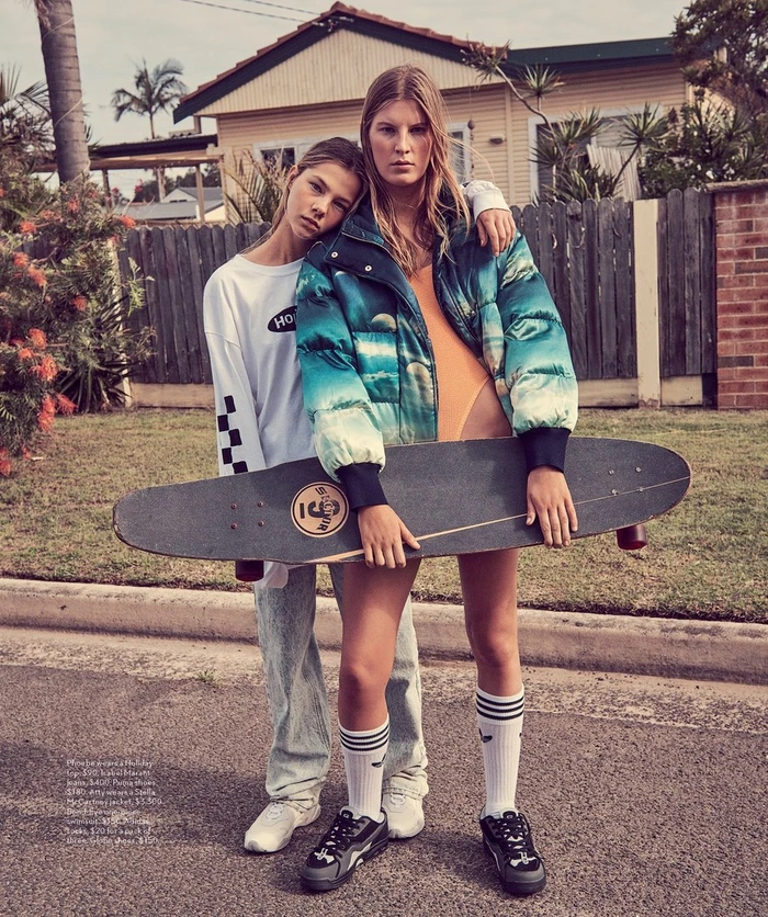 Two young female models posing outdoors holding a longboard skateboard in front of a residential house with a wooden fence.