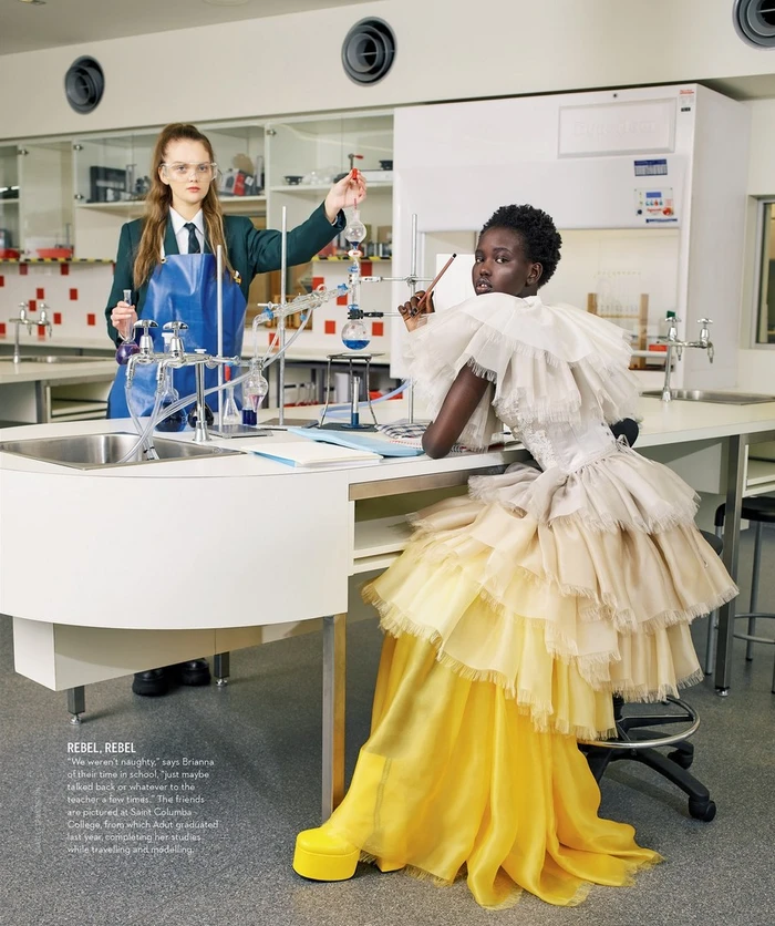 Two models in a science laboratory with one standing holding a flask and one seated in a layered ruffled dress